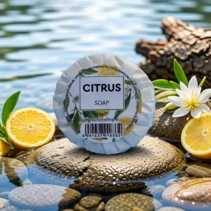 Saponetta rotonda "Citrus Soap" da 15g della linea Hotel Line, posizionata sopra pietre levigate in un ambiente acquatico zen. La confezione plissettata bianca presenta un'etichetta con illustrazioni di limoni e fiori bianchi, circondata da fette di limone fresco e fiori di gelsomino su uno sfondo d'acqua sfocato.