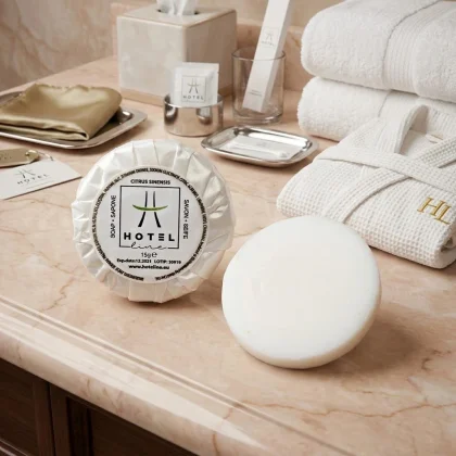 Set cortesia per hotel saponetta plissettata tonda da 15g della linea Hotel Line, presentata su un piano di marmo con asciugamani e accappatoio coordinati.