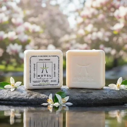 Saponette hotel linea Hotel Line da 15g, una confezionata e una nuda con logo impresso, su pietra bagnata con fiori di zagara e sfondo di alberi in fiore.
