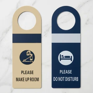 Due cartellini porta hotel in plastica affiancati su marmo: lato oro per la richiesta pulizia camera e lato blu per il messaggio non disturbare con icone.