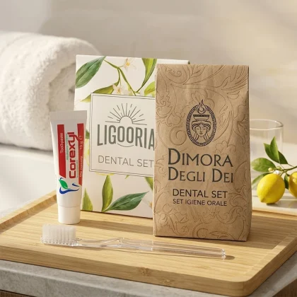 Due varianti di kit igiene orale hotel in sacchetto: versione bianca botanica Ligooria e versione kraft avana Dimora degli Dei con spazzolino Crystal e dentifricio Corexyl su vassoio.