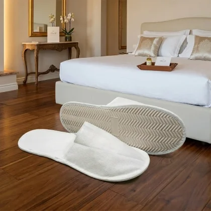 Coppia di pantofole hotel economiche in TNT bianco con suola antiscivolo in gomma EVA, appoggiate sul pavimento in legno di una camera da letto matrimoniale.