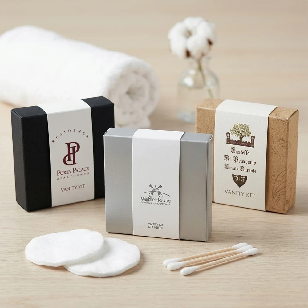 Tre modelli di vanity kit per hotel con fascetta bianca personalizzata su basi nero, grigio satinato e kraft avana con accessori in cotone e legno.