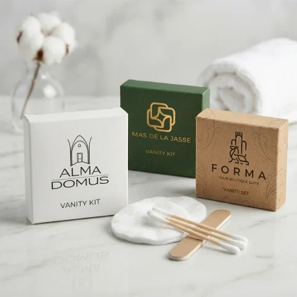 Tre modelli di scatoline per vanity kit hotel: bianco Alma Domus, verde Mas de la Jasse e kraft Forma con accessori in cotone e legno su marmo.